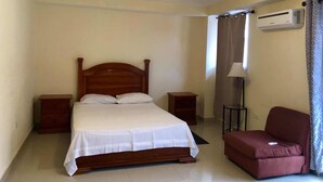 A bedroom with a wooden bed, two bedside tables, a red sofa, a standing lamp, and an air conditioning unit. - FRF Hotel (Cap-Haitien)
