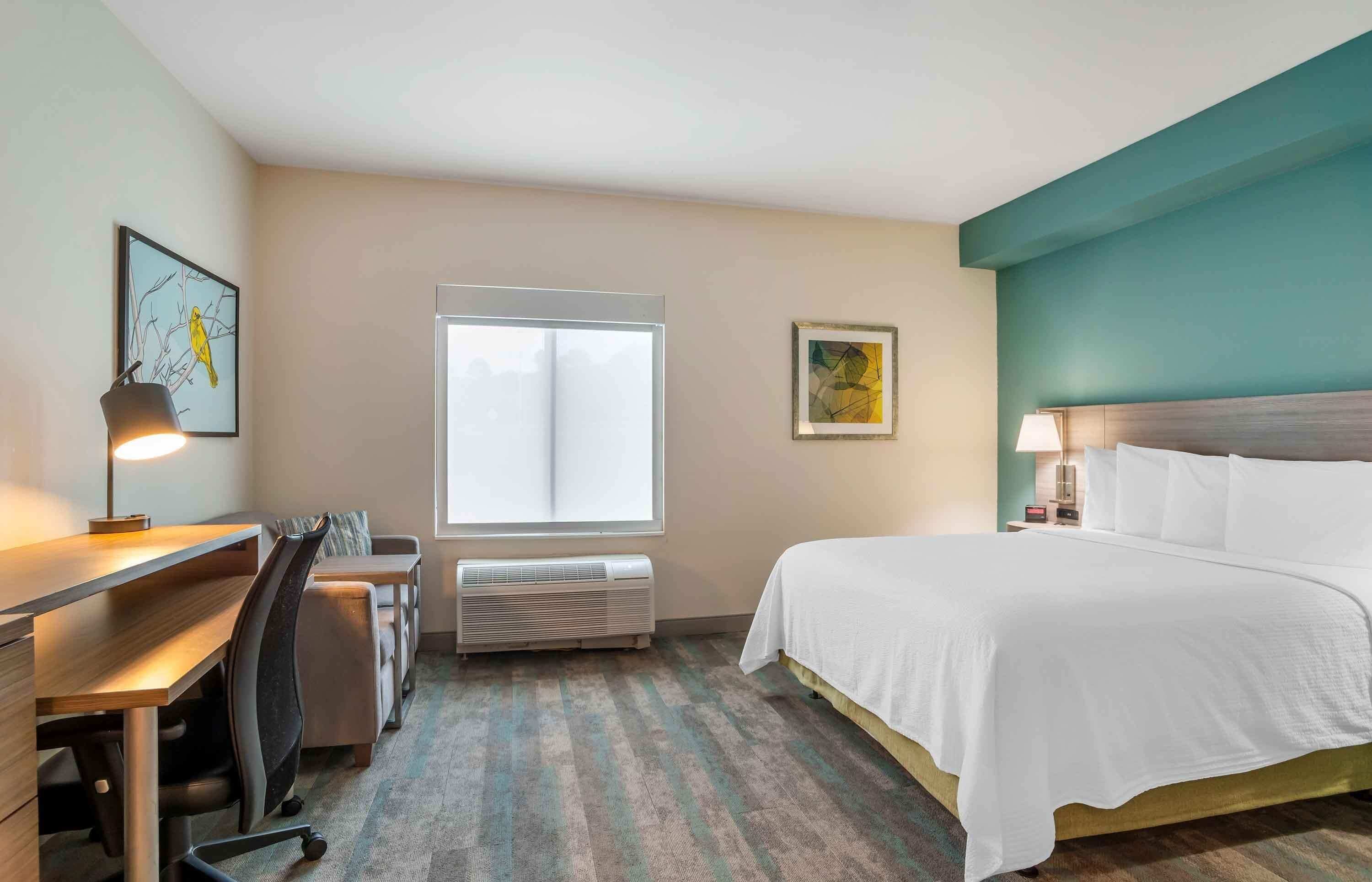 Une chambre d’hôtel équipée d’un lit, d’un bureau, d’une chaise, d’un climatiseur et ornée d’œuvres d’art sur les murs.