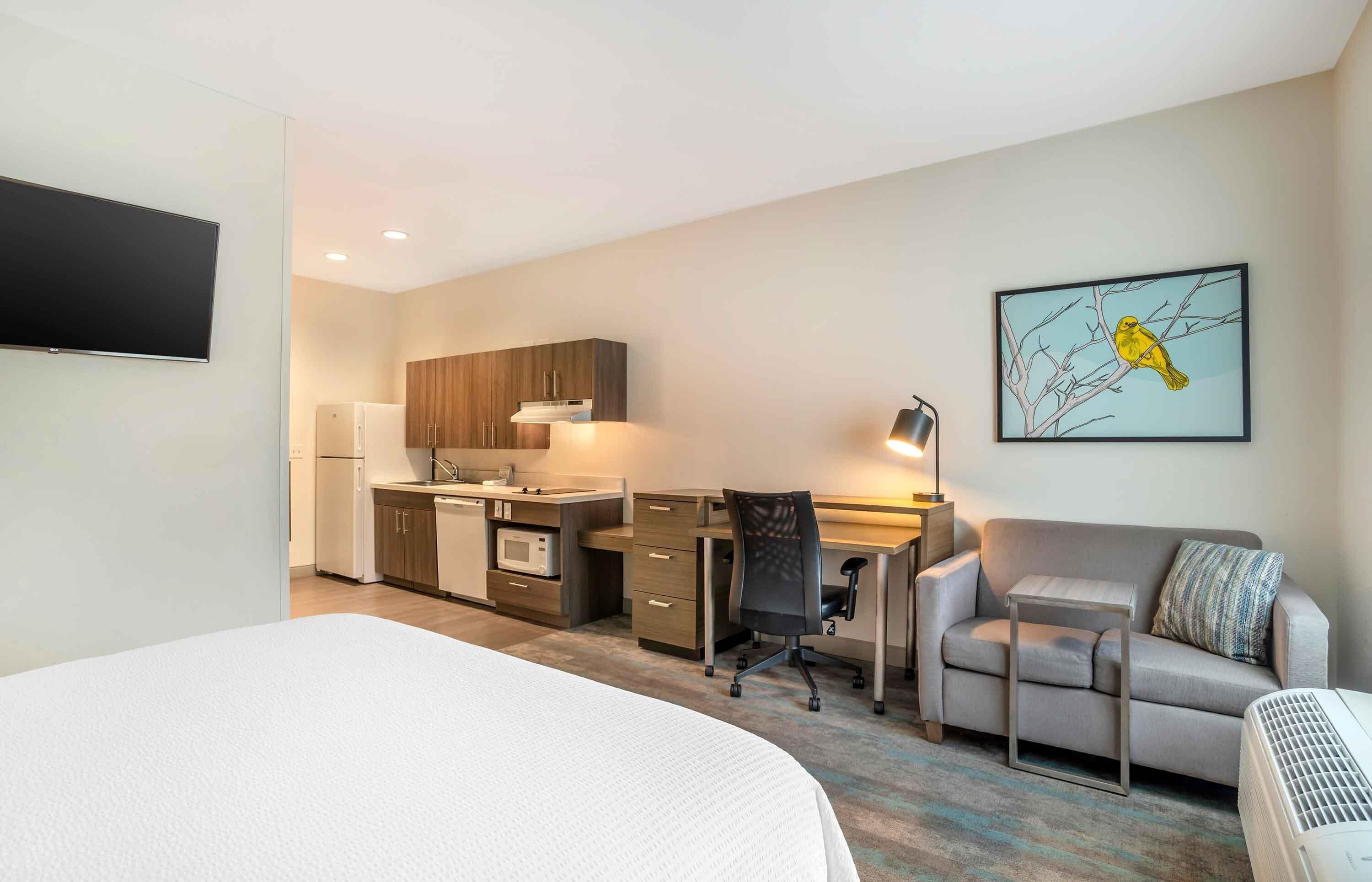 A hotel room with a bed, a desk with a chair, a kitchenette, a sofa, and a wall-mounted TV.