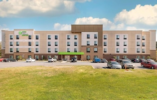 Un hotel de varios pisos con fachada plana, entrada verde y estacionamiento con varios autos.