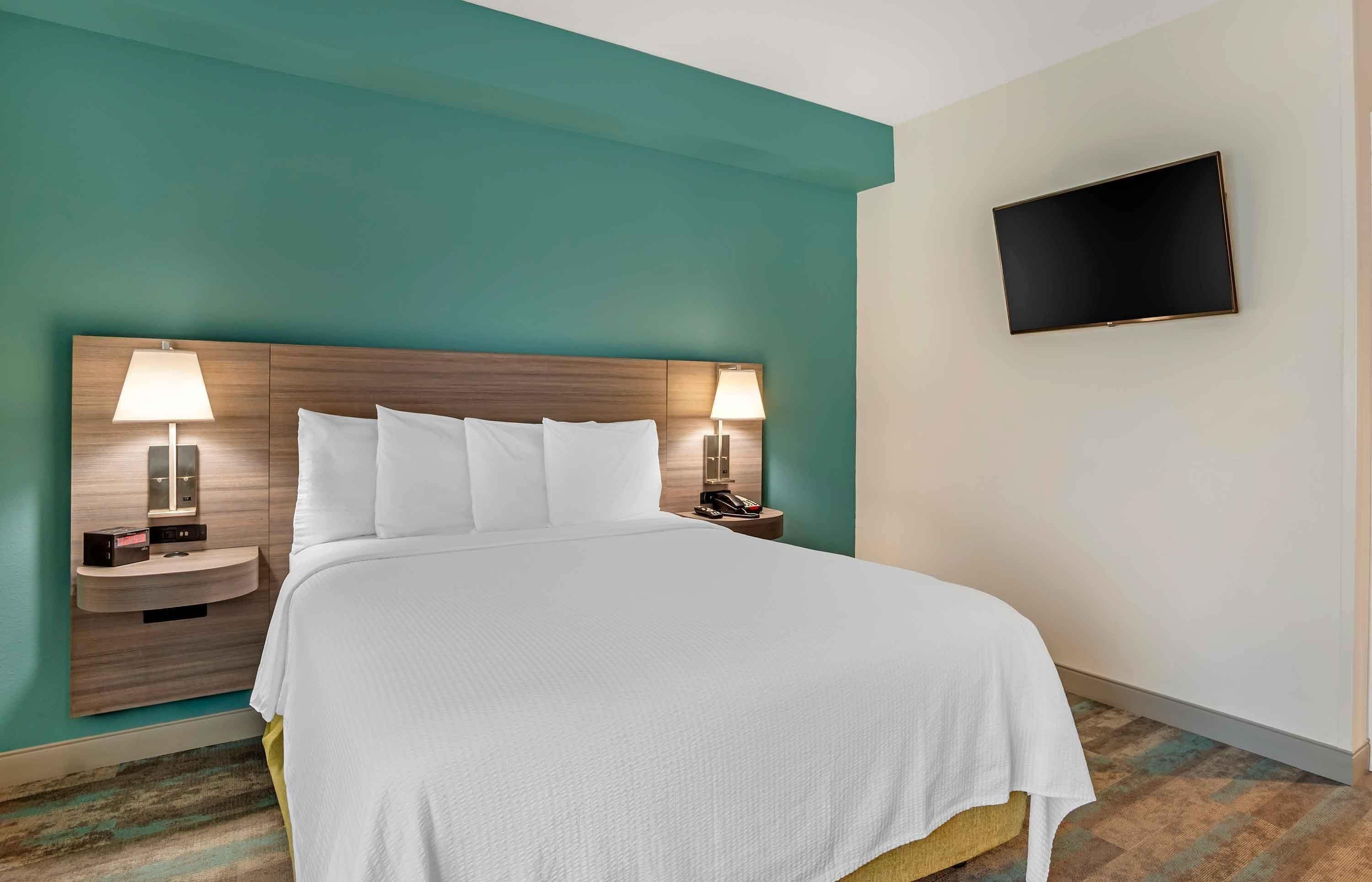A hotel room with a bed, two bedside tables with lamps, a flat-screen TV mounted on the wall, and a teal-colored wall.