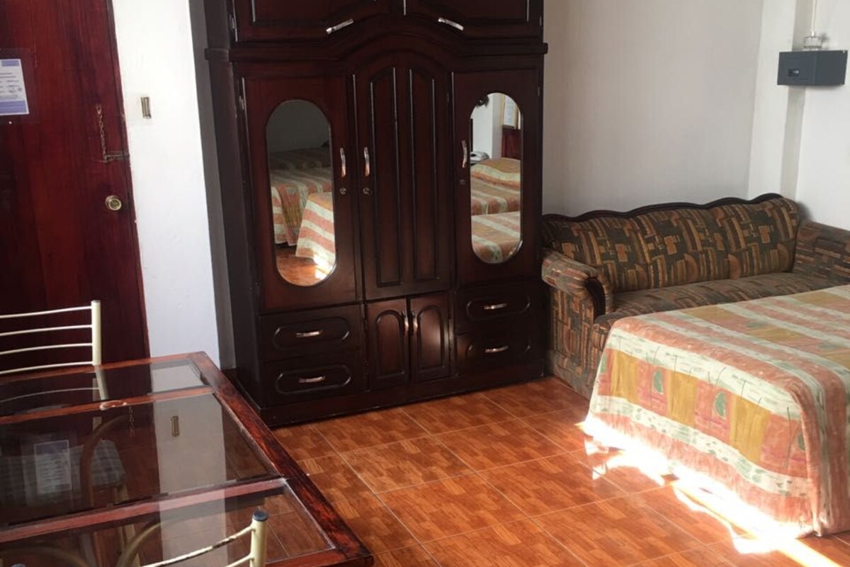 Een kamer met een houten garderobe, een bed met een bedovertrek met patroon, een eettafel met stoelen en een betegelde vloer.