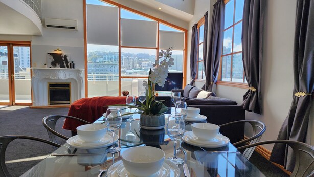 A dining area with a glass table set for two, a fireplace, and a city view through large windows. - U Suites on Courtenay (Wellington)