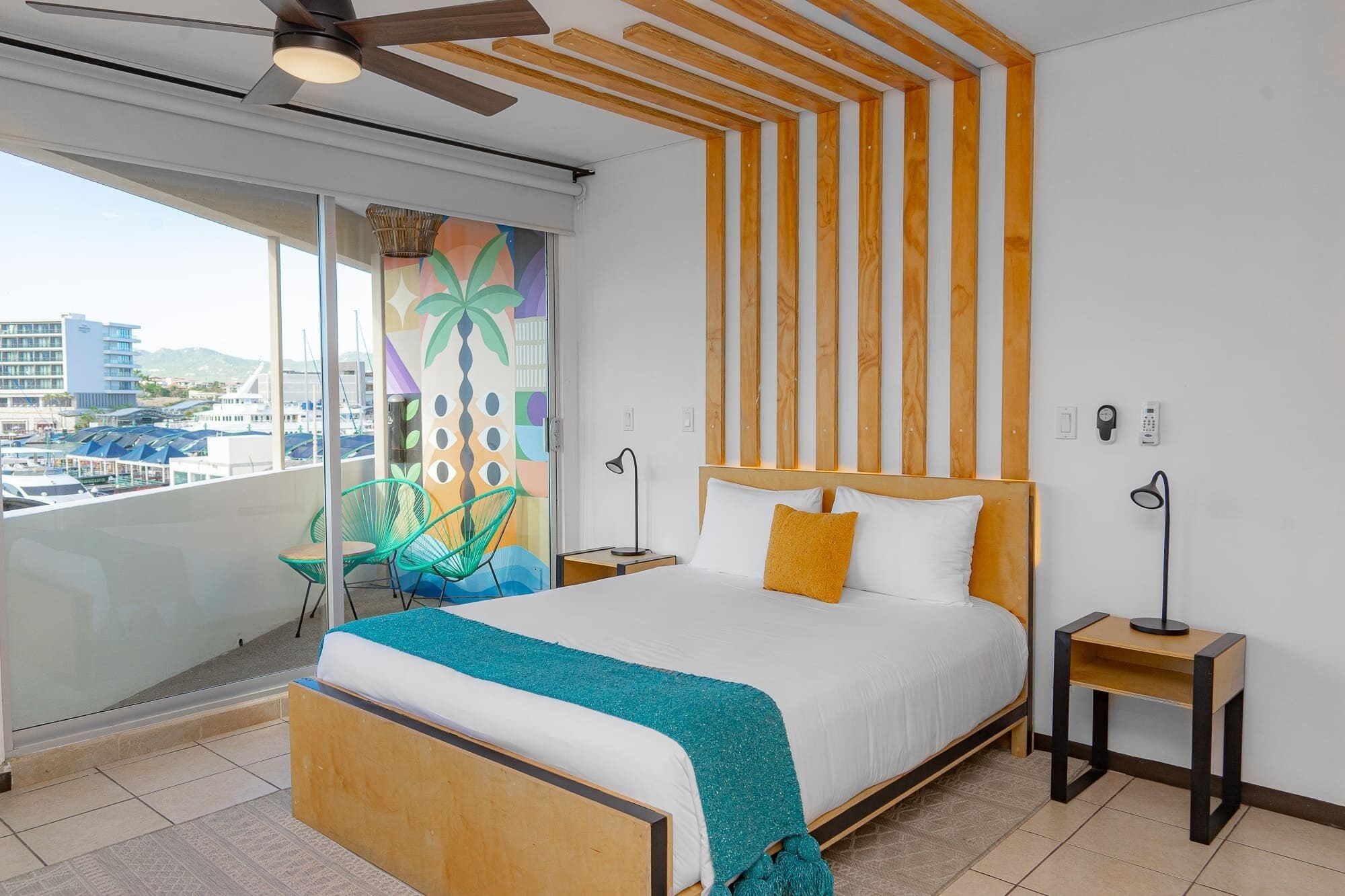 A hotel room with a bed, a nightstand, a chair, a window with a view of the city, and a ceiling fan.