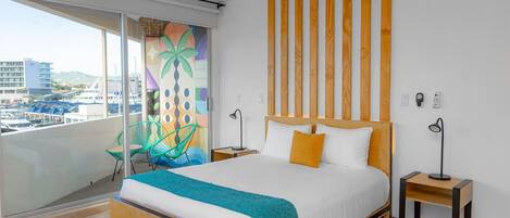 A hotel room with a bed, a nightstand, a chair, a window with a view of the city, and a ceiling fan.