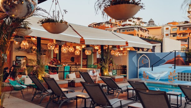 An outdoor seating area with lounge chairs and umbrellas, a pool with a mural, and a bar area with plants and hanging lights. - Mayan Monkey Los Cabos | Social Hotel (Cabo San Lucas)