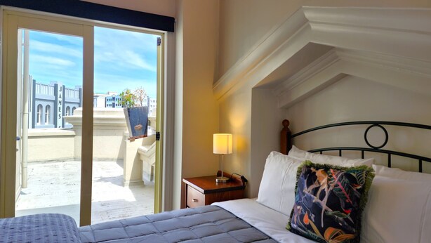 A hotel room with a bed, a nightstand, a lamp, a window with a view of buildings, and a balcony with a potted plant. - U Suites on Manners (Wellington)