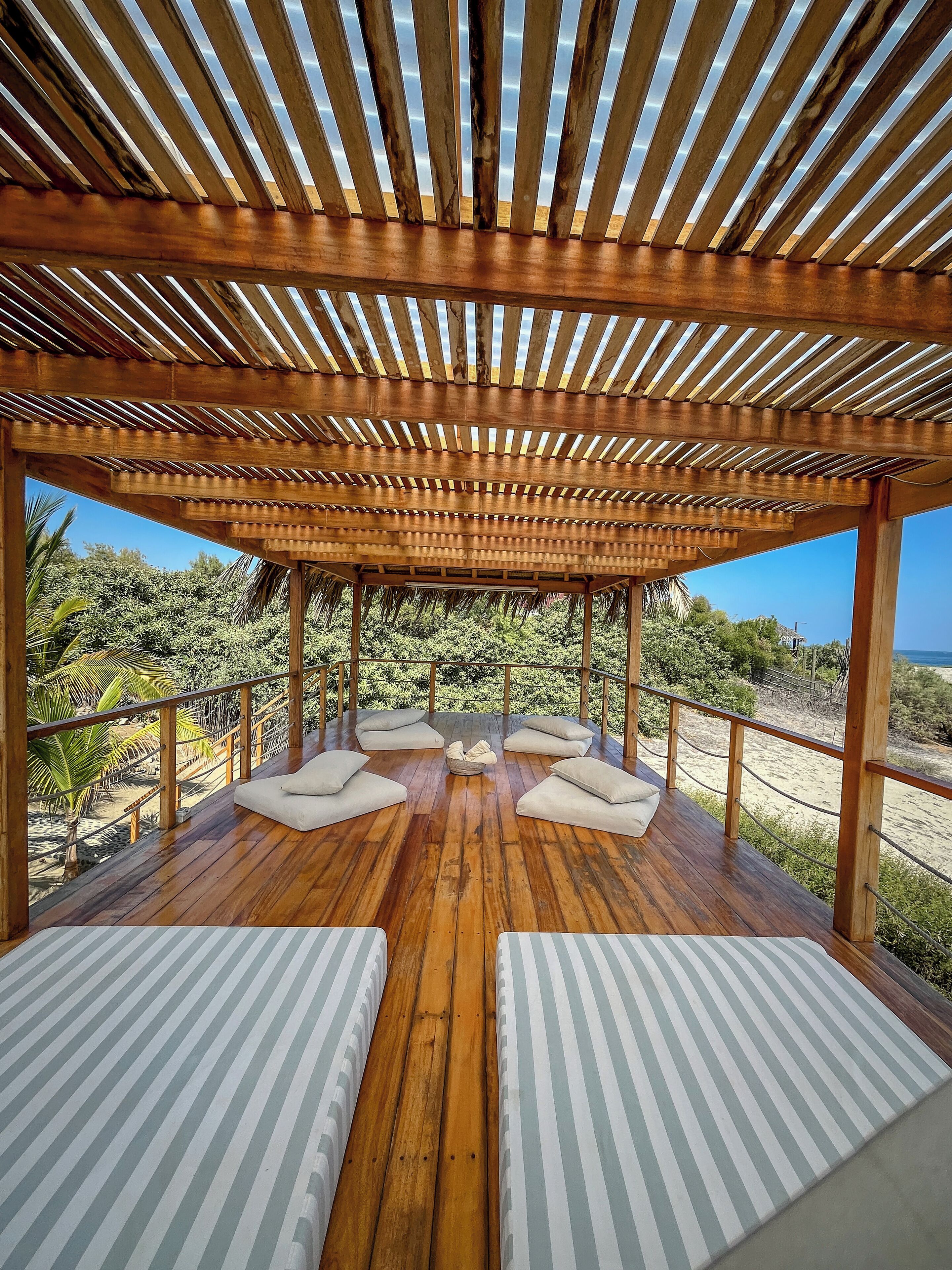A wooden deck with a pergola, white cushioned seating, and a view of the beach.