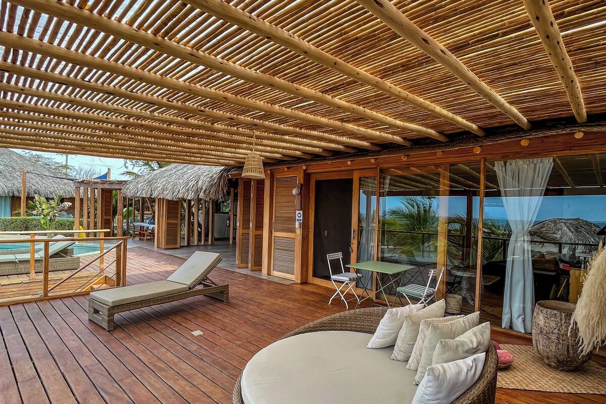 A wooden deck with a thatched roof, a wicker sofa, and a pool area.