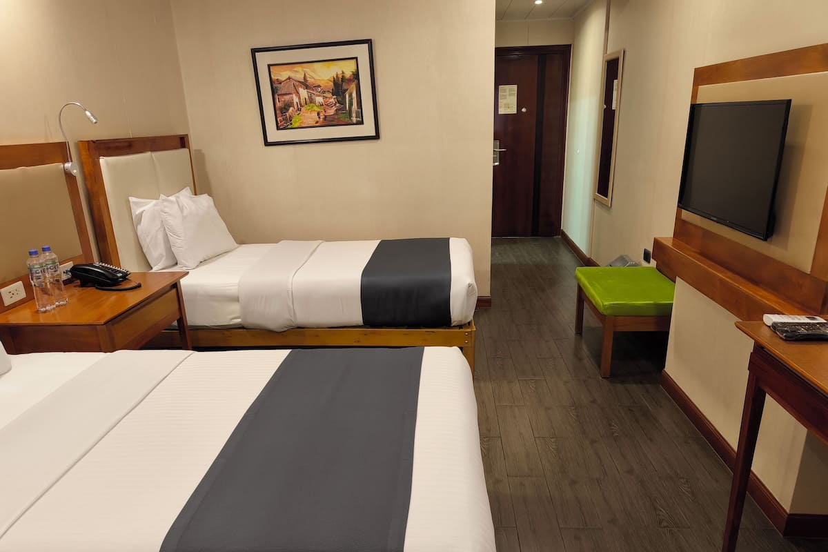 Ein Hotelzimmer mit zwei Betten, einem Schreibtisch mit Stuhl, einem Fernseher, einem Bild an der Wand und Holzboden.