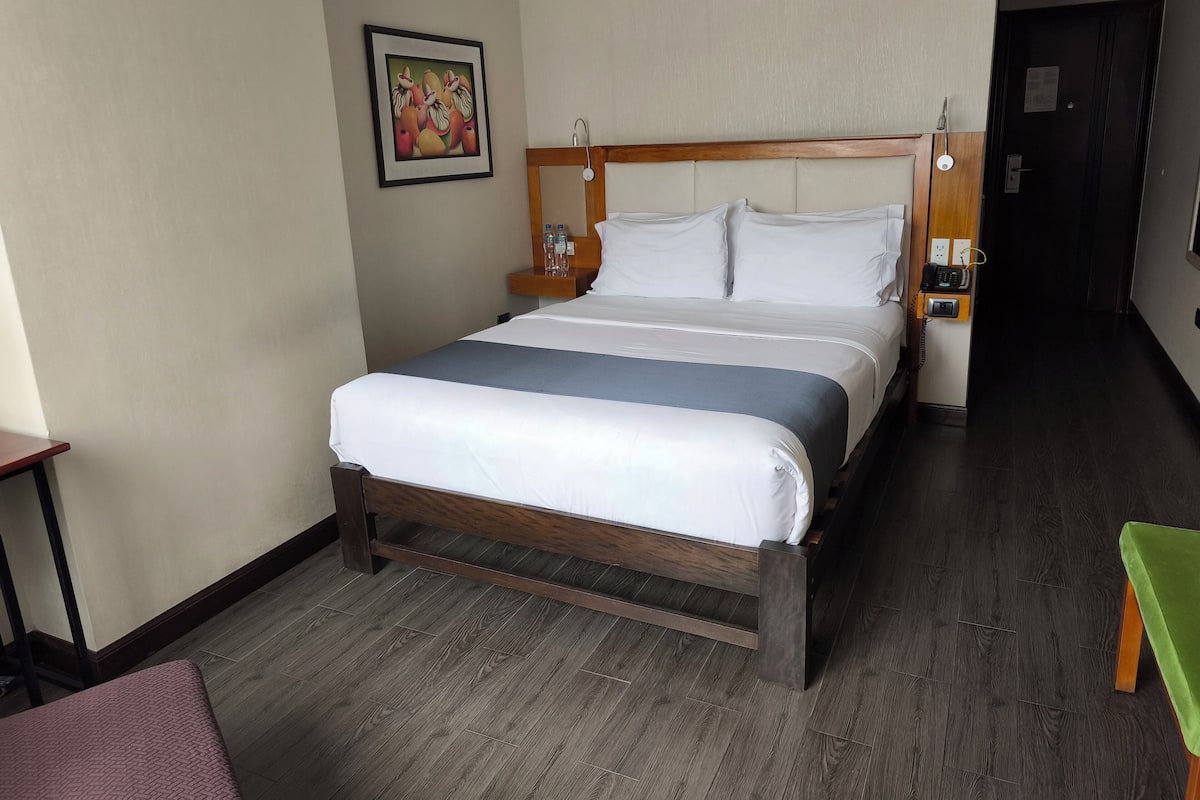 Ein Hotelzimmer mit einem hölzernen Bett, weißer Bettwäsche, einem Nachttisch mit Lampe, einem fest montierten Telefon und einem gerahmten Bild.