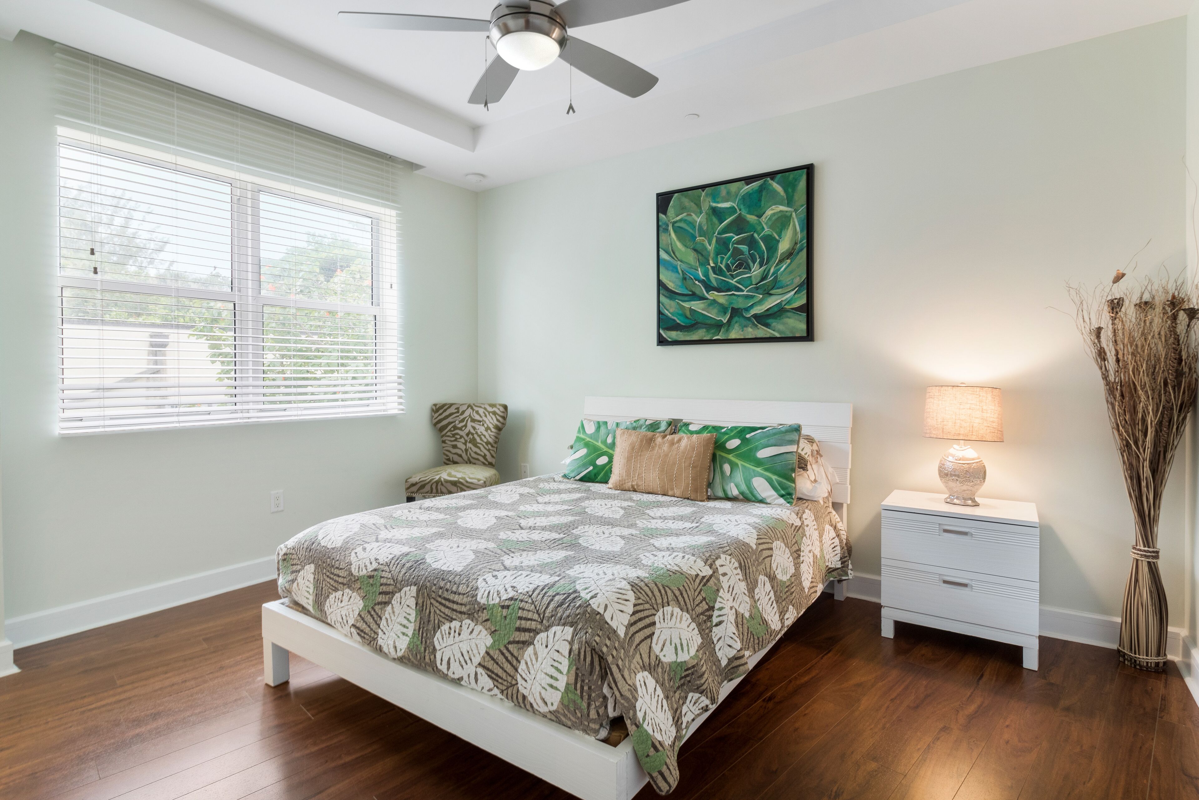 A bedroom with a bed, nightstands, a ceiling fan, a window with blinds, and a wall-mounted painting.