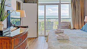 Quarto de hotel com uma cama, cômoda de madeira, televisão e vista para o oceano através de uma grande janela.