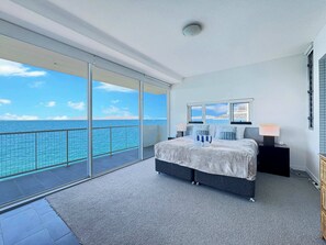 A bedroom with a large bed, a nightstand, a lamp, and a view of the sea through large windows.