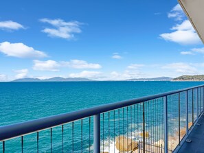 Une vue dégagée sur la mer depuis un balcon doté d’une rambarde en métal, avec, au loin, une terre ferme et des montagnes, le tout sous un ciel bleu.