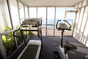 Ein Fitnessraum mit Laufbändern und Ergometer, große Fenster mit Meerblick und ein Balkon mit Pflanzen.