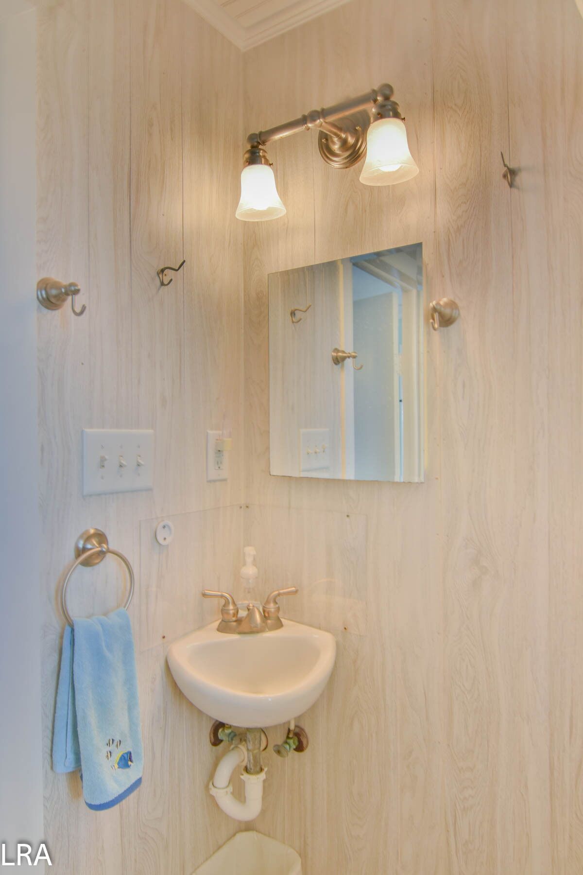 Ein Badezimmer mit einer Holzwand, einem weißen Waschbecken mit goldener Armatur, einem Handtuchhalter mit blauem Handtuch und einem Spiegel mit Leuchte darüber.