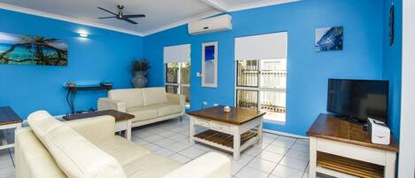 A living room with a sofa, coffee table, TV stand, ceiling fan, and large windows.