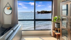 A room with a large window offering a view of the sea, a sofa, a cabinet, and a mirror on the wall. - Blue on Cockle (Cockle Bay)