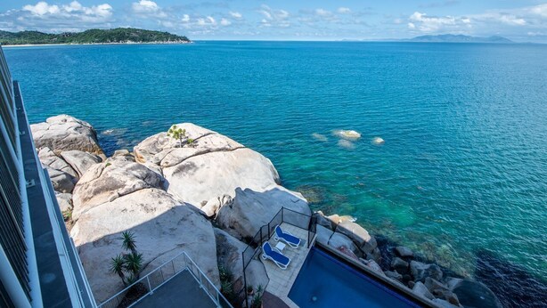 A pool area with lounge chairs, a pool, and a rocky shoreline with clear blue water.