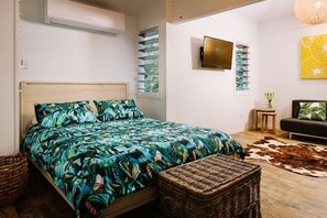 A bedroom with a bed, a wicker chair, a cowhide rug, a small table, and a wall-mounted TV. - The Little Bush Hut (Nelly Bay)