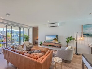 A modern living room with a large window, a flat-screen TV, leather sofas, and a balcony with a city view. - Blue on Blue Apartment 1011 (Nelly Bay)