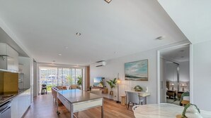 Una cocina moderna con una isla central, una zona de comedor con mesa redonda y un espacio de sala de estar con televisor y plantas.