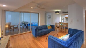 A modern living room with a blue sofa, wooden flooring, a dining area, and a kitchen in the background. - Maggies Beachfront Apt. 4 (Horseshoe Bay)