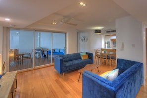 A modern living room with a blue sofa, wooden flooring, a dining area, and a kitchen in the background.