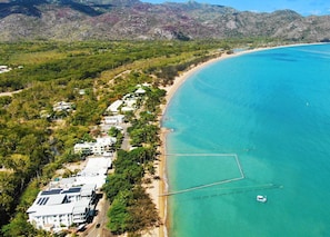 An aerial view of a coastal area with a beach, buildings, and clear blue water.