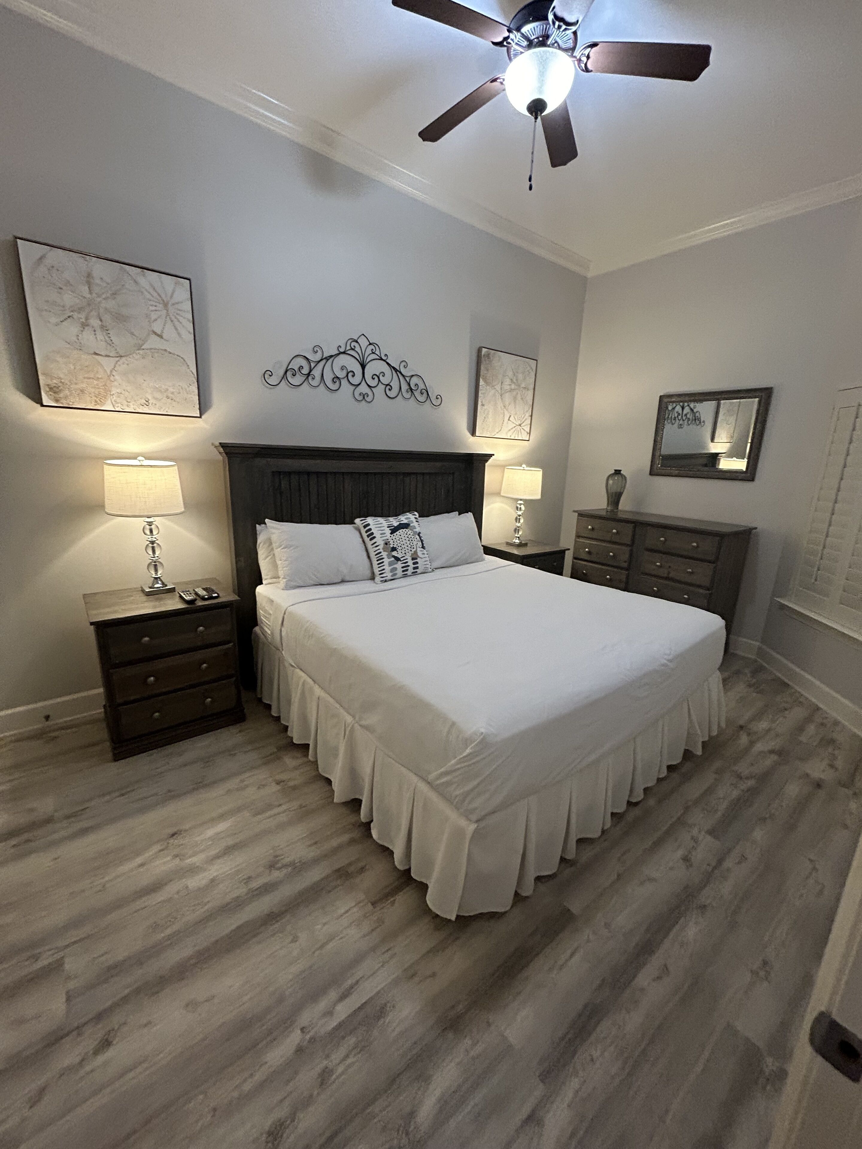 A bedroom with a large bed, wooden floor, two bedside tables with lamps, a ceiling fan, and artwork on the walls.
