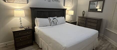 A bedroom with a large bed, wooden floor, two bedside tables with lamps, a ceiling fan, and artwork on the walls.