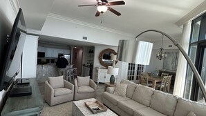 Amplia sala de estar con un sofá modular grande, una mesa de centro y un televisor sobre un mueble bajo. Se aprecia un ventilador de techo, una araña y vistas al área de cocina y comedor.