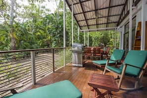 Eine überdachte Terrasse im Freien mit Holzmöbeln, einem Grill und Blick auf üppiges Grün.