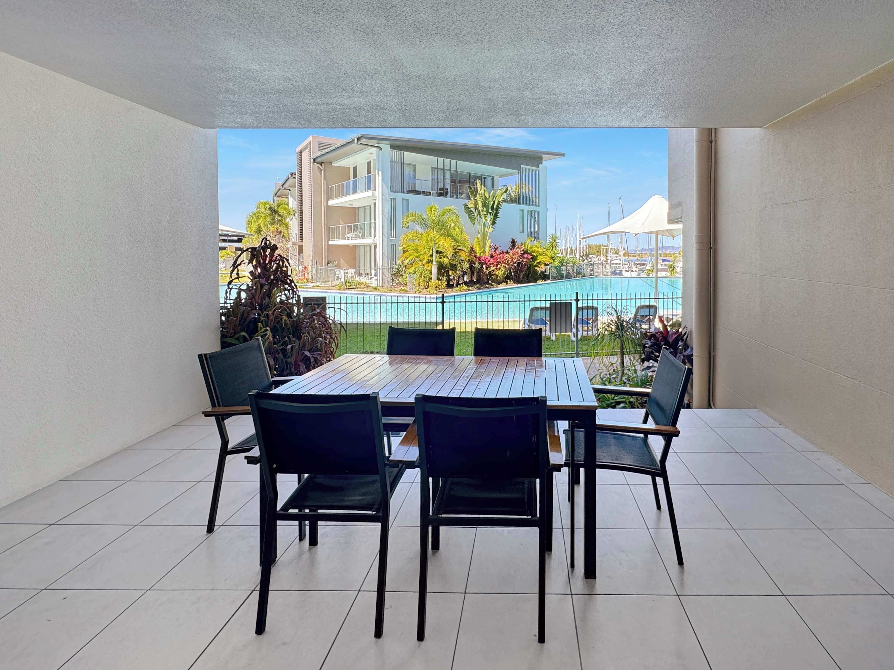 A balcony with a dining table and chairs, overlooking a swimming pool and a building.