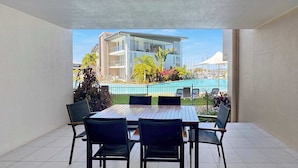 A balcony with a dining table and chairs, overlooking a swimming pool and a building. - Blue on Blue Apartment 813 (Nelly Bay)