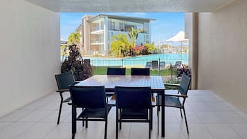 A balcony with a dining table and chairs, overlooking a swimming pool and a building.