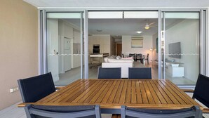 A dining area with a wooden table and chairs, a kitchen in the background, and a sliding glass door leading to a living space. - Blue on Blue Apartment 813 (Nelly Bay)