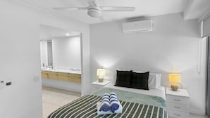 A hotel room with a bed, two bedside tables with lamps, a ceiling fan, a bathroom visible through an open door, and a large mirror. - Blue on Blue Apartment 813 (Nelly Bay)