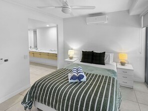 Quarto de hotel com cama, duas mesas de cabeceira com abajures, ventilador de teto, banheiro visível por uma porta aberta e um espelho grande.