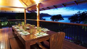 A wooden deck with a table set for dining, overlooking a body of water and mountains.