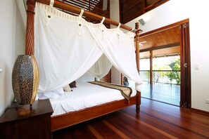 A wooden four-poster bed with white bedding and a canopy, a decorative vase on a bedside table, and a view of an outdoor balcony. - Villa Kembali (Horseshoe Bay)