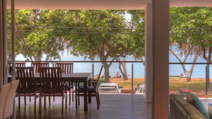 Una habitación con una mesa y sillas de madera, un sofá y vistas a la playa, con árboles y gente.