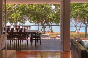 A room with a wooden table and chairs, a sofa, and a view of the beach with trees and people.