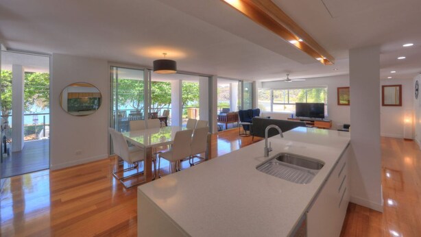 Una cocina moderna con una isla central, un comedor con sillas, un televisor y un ventanal con vistas a árboles.