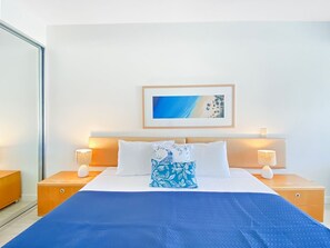 A neatly arranged hotel room with a bed, two bedside tables, a nightstand, a wall-mounted lamp, and a framed picture above the bed.