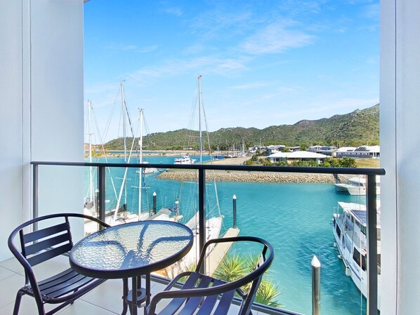 Un balcon avec une table et des chaises donnant sur une marina où sont amarrés des bateaux, et sur un paysage vallonné.