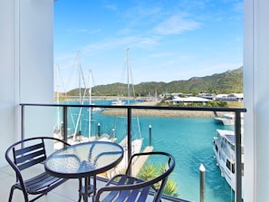 A balcony with a table and chairs overlooking a marina with boats and a hilly landscape.