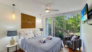 A bedroom with a bed, a ceiling fan, a TV, and a view of greenery outside. - Maggies Beachfront Apt. 2 (Horseshoe Bay)