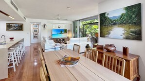 A modern kitchen with a wooden dining table, white bar stools, and a large window with a view of greenery. - Maggies Beachfront Apt. 2 (Horseshoe Bay)